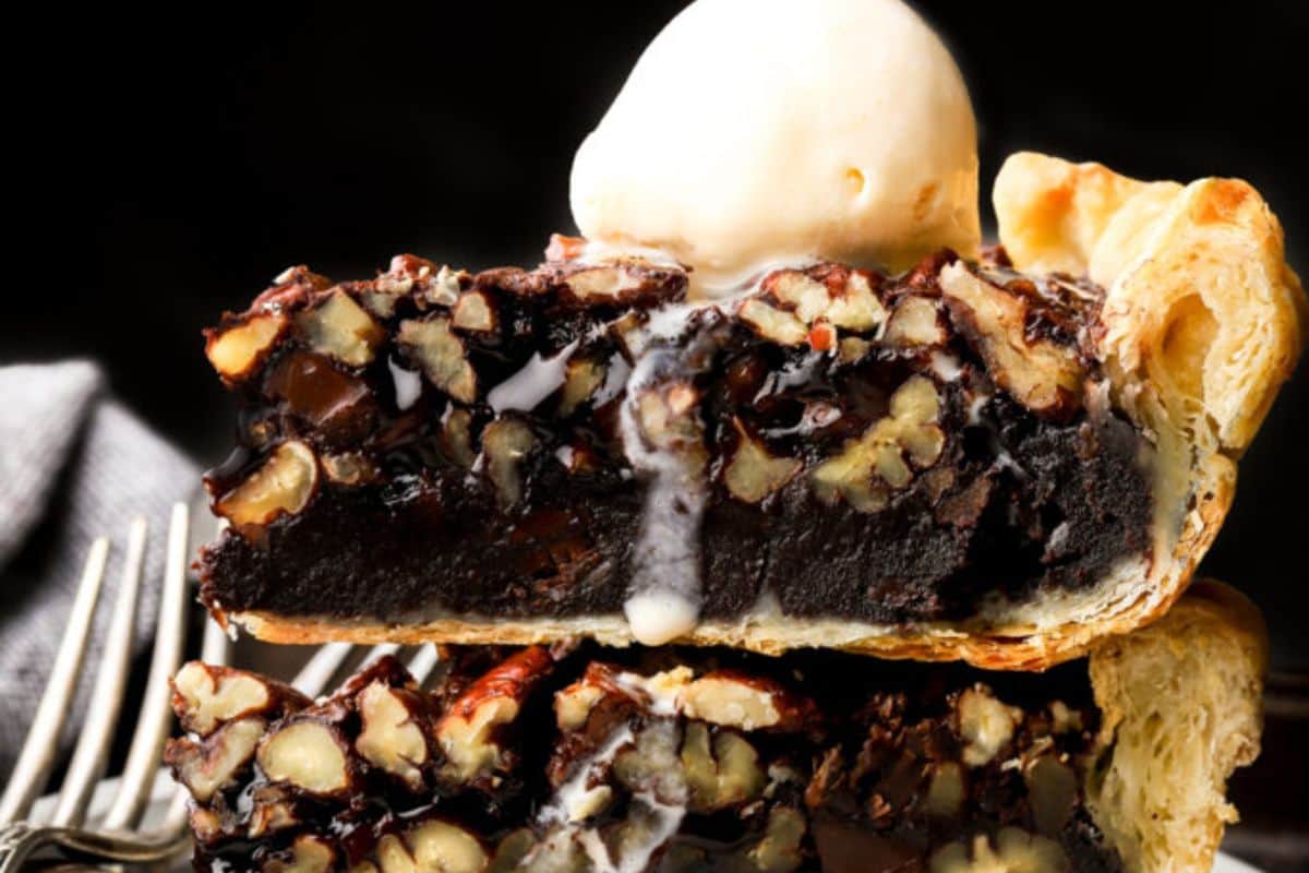 Close-up of a slice of pecan chocolate pie topped with a scoop of vanilla ice cream. The pie has a flaky crust, rich chocolate filling, and a generous amount of pecans. The melting ice cream adds a touch of creaminess to the decadent dessert.