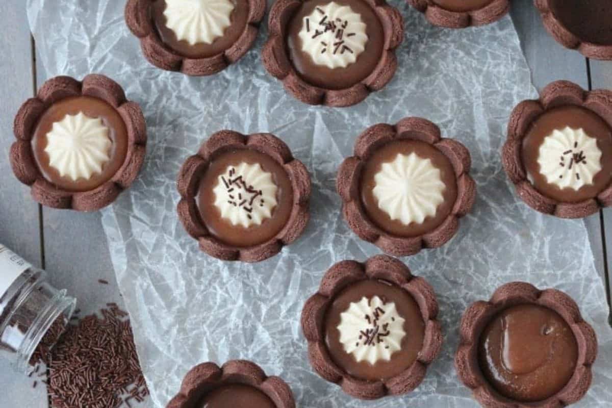 Top view of several chocolate tarts with different toppings arranged on a crinkled sheet of parchment paper. Some tarts have white icing, some have chocolate sprinkles, and others are plain. A small jar with chocolate sprinkles is seen on the left.