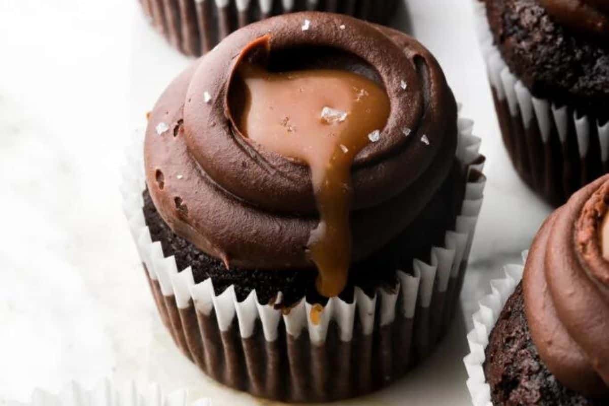 A close-up of a chocolate cupcake with creamy chocolate frosting topped with a drizzle of caramel sauce that's slightly dripping down the side. The cupcake is in a white paper liner, and there are more similar cupcakes in the background.