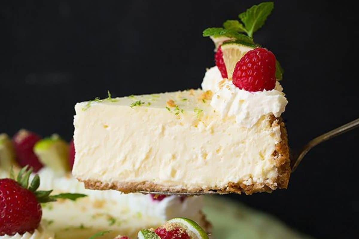 A close-up of a slice of creamy cheesecake being lifted out of a larger cheesecake. The slice is topped with whipped cream, a fresh raspberry, a small slice of lime, and a mint leaf. The cheesecake has a graham cracker crust and is garnished with similar toppings.