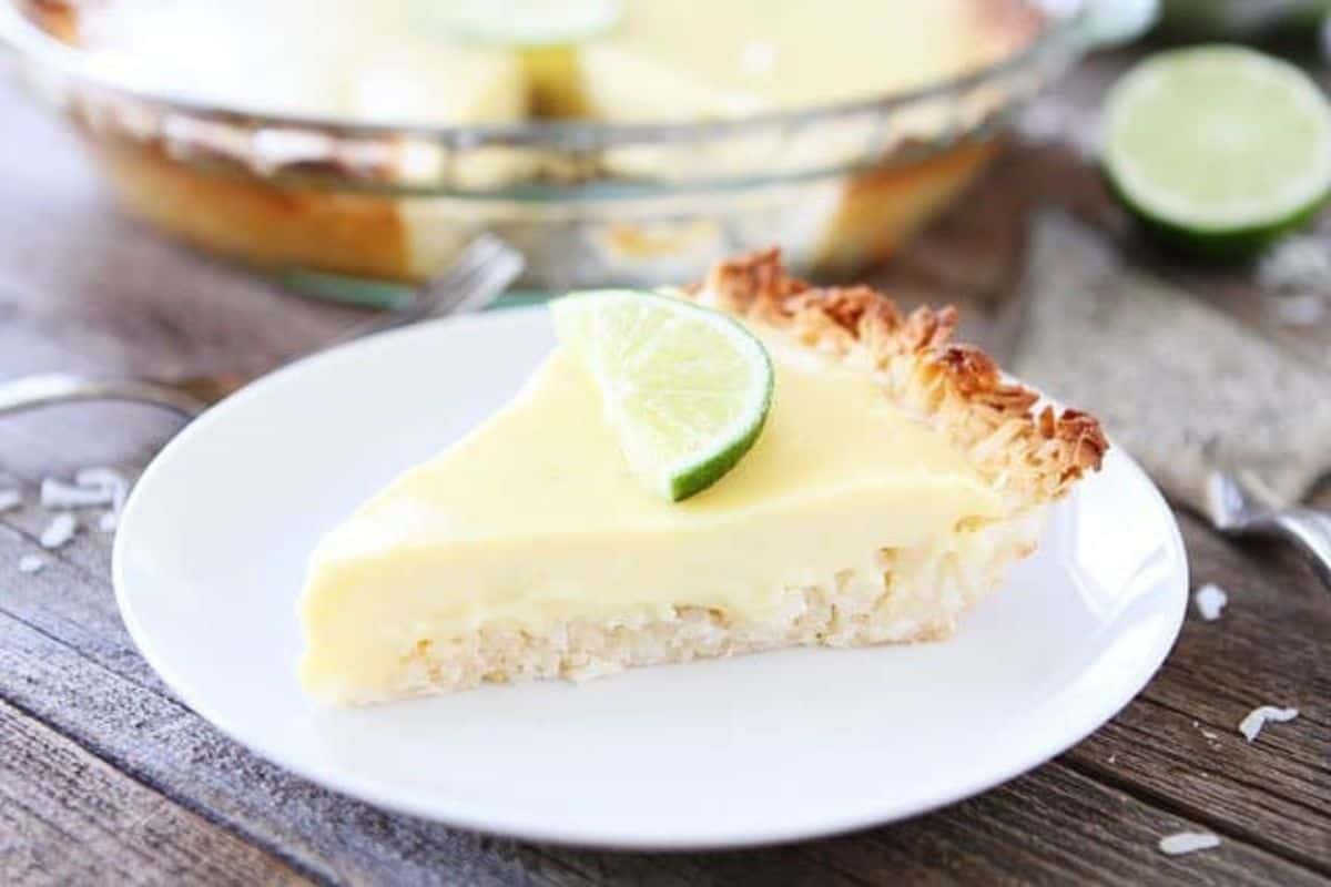 A slice of creamy lime pie garnished with a lime wedge sits on a white plate. The pie has a toasted coconut crust and a smooth, pale yellow filling. In the background, the full pie is partially visible on a wooden surface.