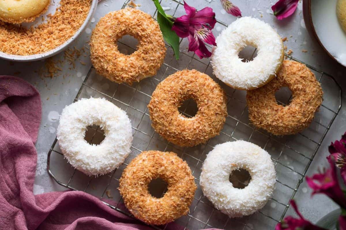 A cooling rack with seven baked coconut-covered donuts. Four donuts are coated in toasted coconut, while three are covered in white coconut flakes. Surrounding the rack are pink flowers, a pink cloth, and a bowl partially visible with more donuts.