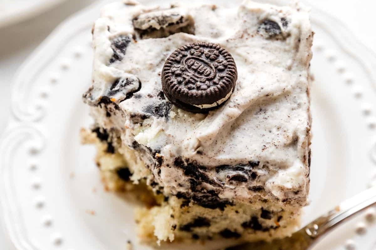 A close-up of a slice of cookies and cream cake on a white plate. The cake has a white and black speckled frosting with bits of crushed cookies mixed in. An uncrushed chocolate sandwich cookie is placed on top as decoration. A silver fork is partially visible.