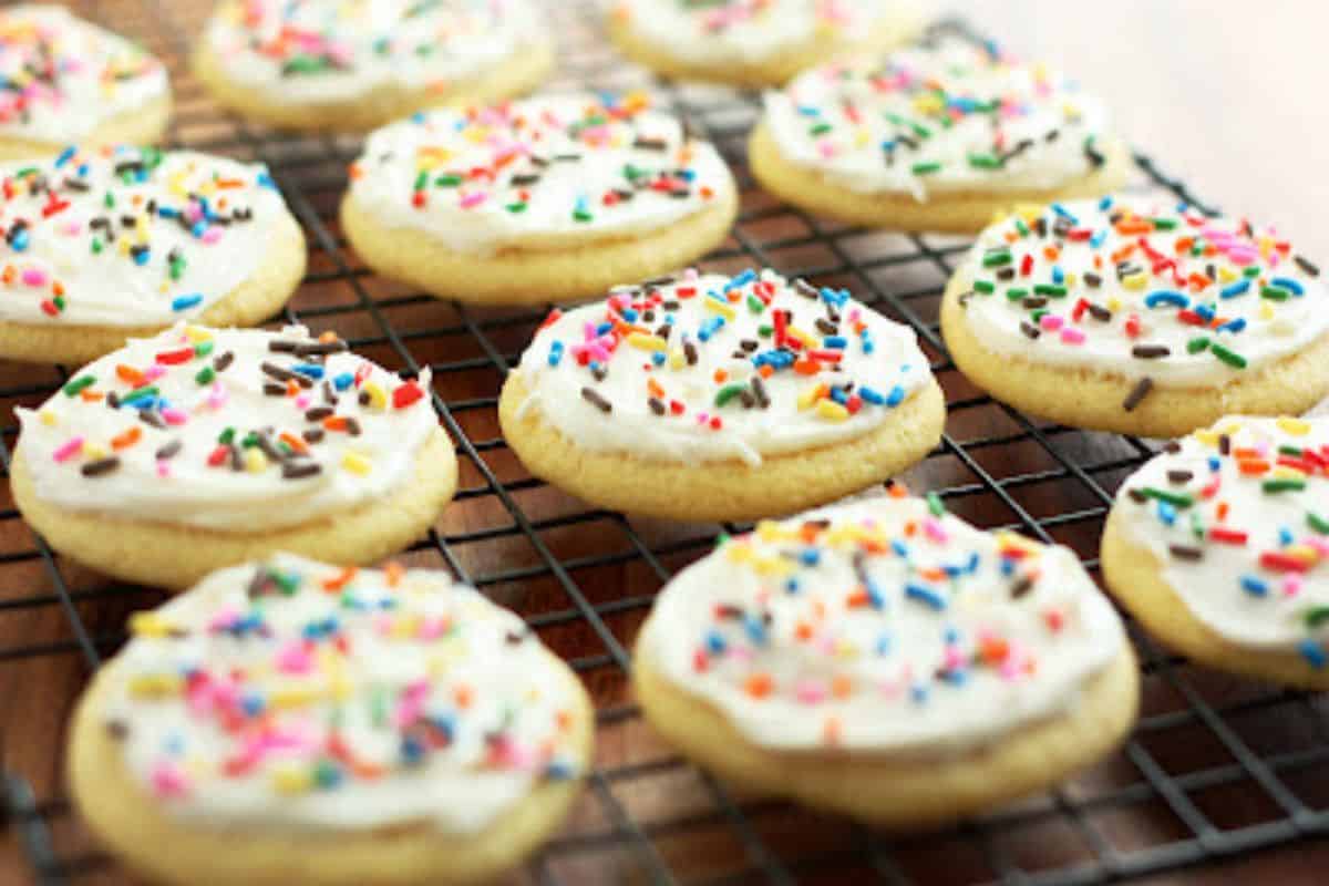 Several sugar cookies are cooling on a black wire rack. Each cookie is topped with a layer of white frosting and colorful sprinkles. The cookies are neatly arranged in rows and are set against a blurred wooden background.