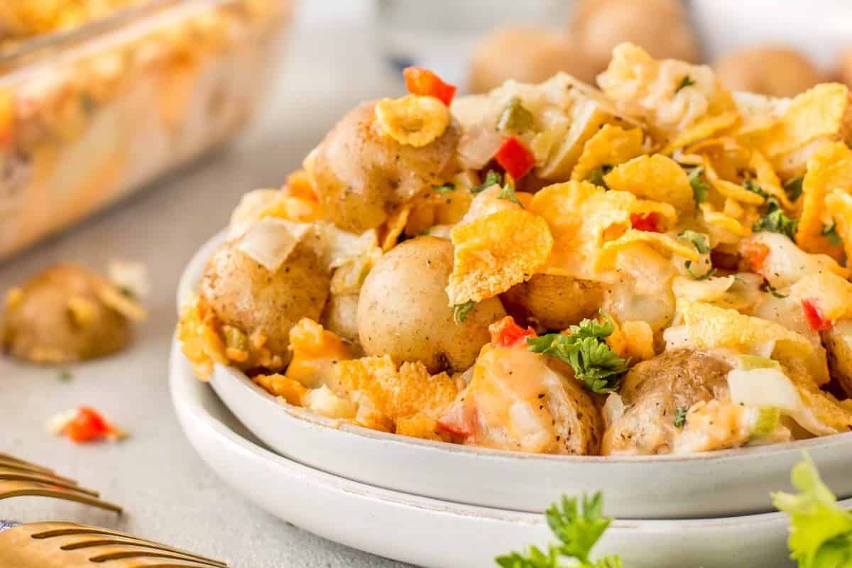 A close-up of a bowl filled with a creamy potato casserole. The dish contains small whole potatoes, shredded cheese, diced red and green peppers, and is topped with crumbled potato chips. A garnish of fresh parsley adds a touch of color, with a similar dish in the background.