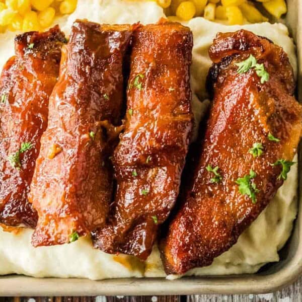 Four pieces of glazed pork belly atop a bed of creamy mashed potatoes, accompanied by corn kernels.