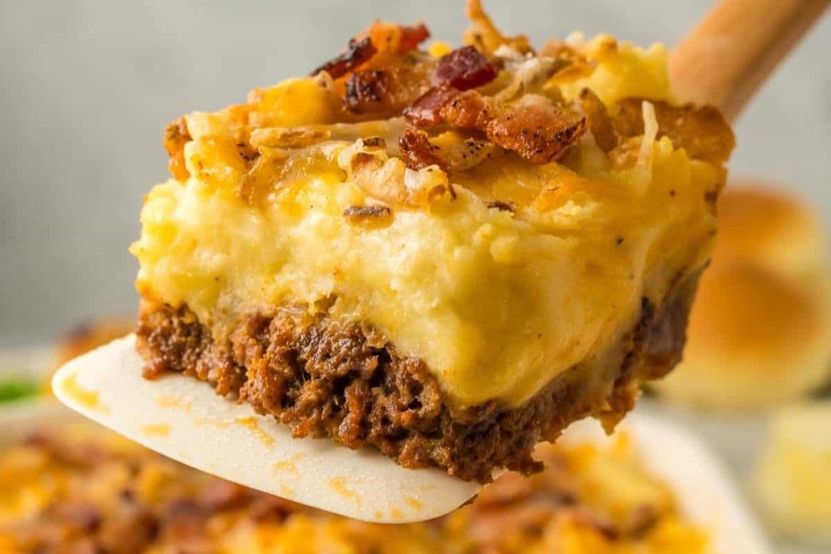 A close-up of a serving of shepherd's pie being lifted with a spatula. The piece has layers of minced meat, creamy mashed potatoes, and is topped with melted cheese and crispy bacon bits. The background shows more of the dish and some dinner rolls.