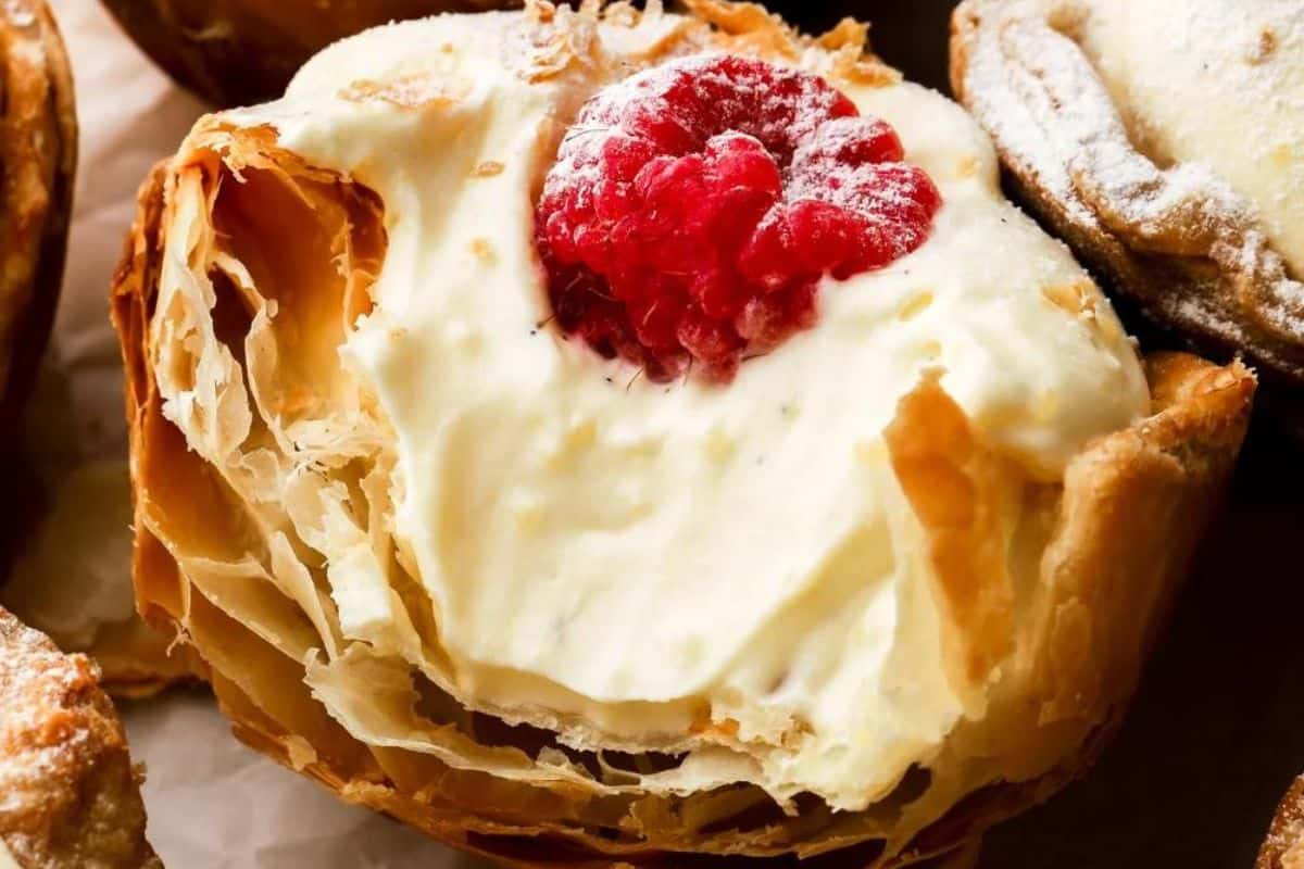 A close-up of a flaky pastry filled with creamy, whipped filling and topped with a fresh red raspberry. The pastry's layers are golden brown and crispy, and a light dusting of powdered sugar is visible on a nearby pastry.