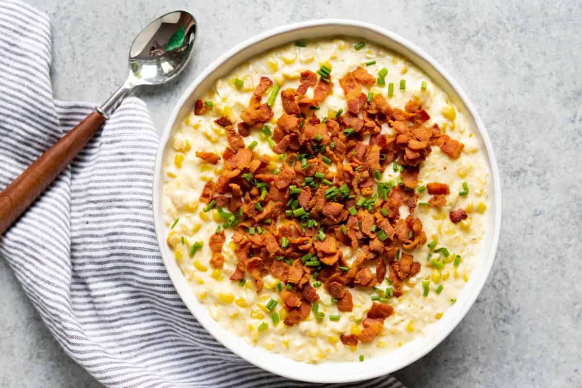 A bowl filled with creamy corn casserole topped with crispy bacon bits and chopped chives, placed on a light gray surface. A white and gray striped cloth is next to the bowl, with a spoon resting on it.