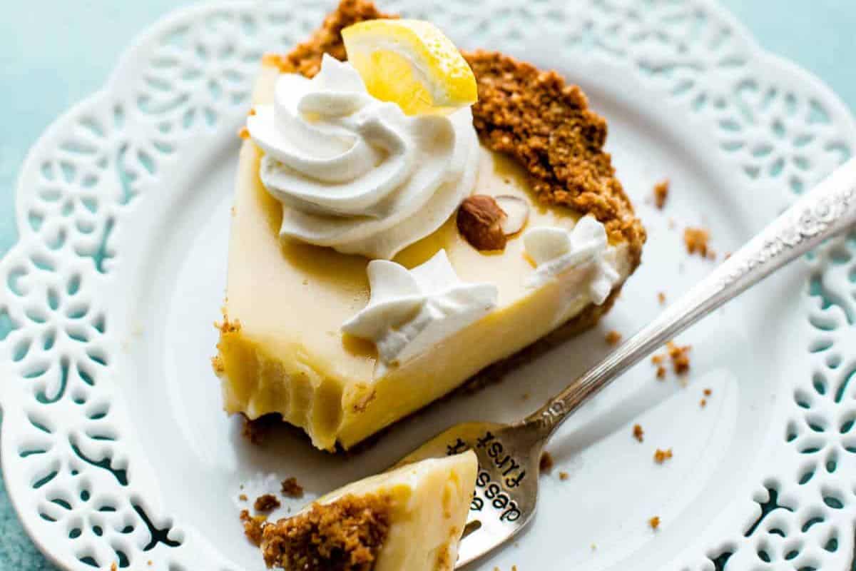 A slice of lemon pie on a lace-edged plate, garnished with a dollop of whipped cream and a lemon wedge. A fork rests nearby with a small piece of pie, and the pie has a crumbly crust.