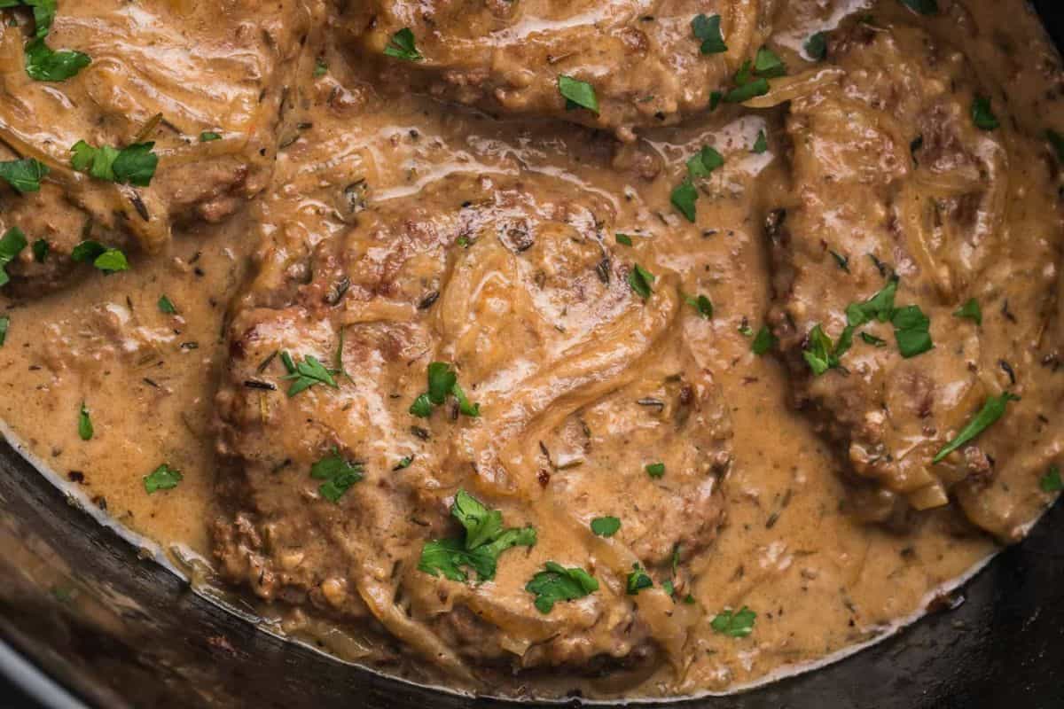 Four beef patties in a creamy brown gravy, garnished with chopped parsley.
