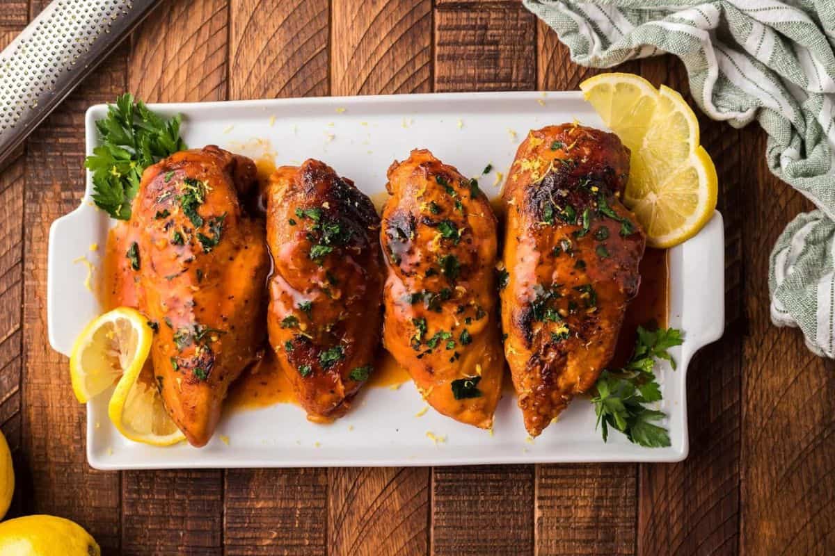 Four glazed chicken breasts garnished with chopped herbs and lemon slices, presented on a rectangular white plate. The plate is set on a wooden surface with a green-striped cloth napkin and additional lemon slices nearby.