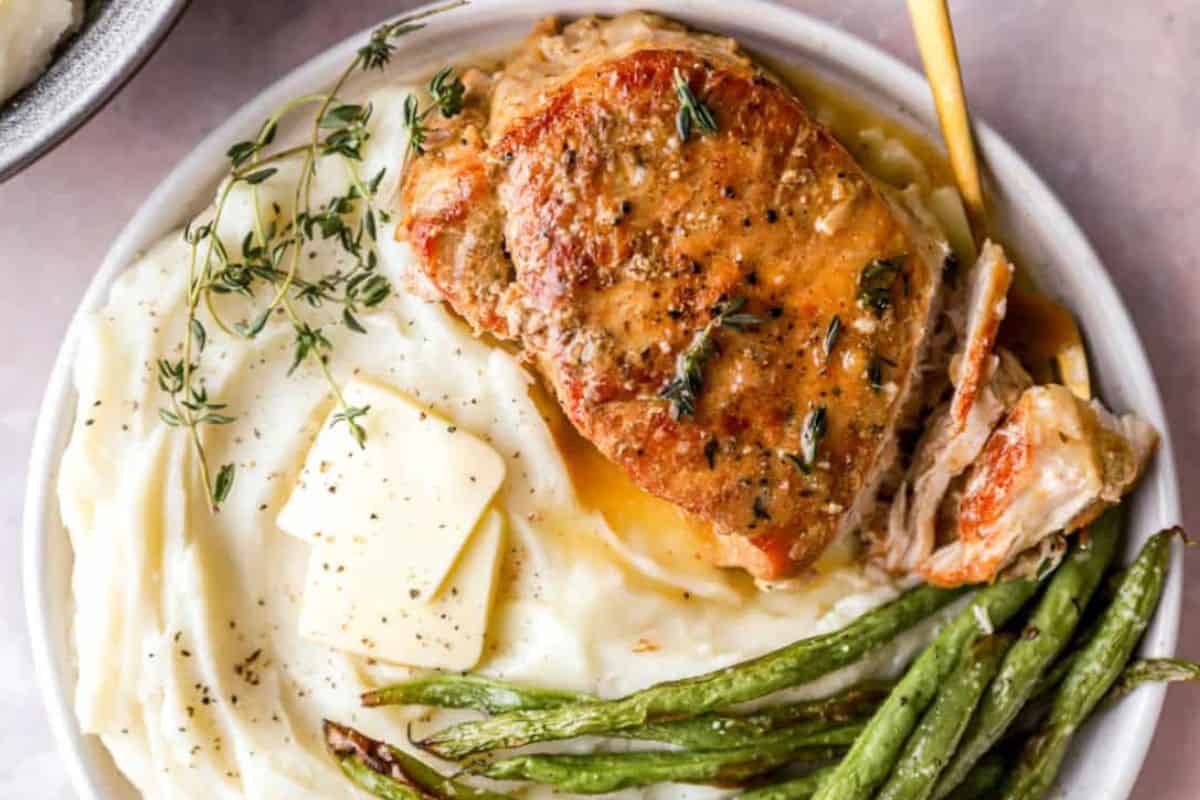 A plate of creamy mashed potatoes topped with butter and fresh herbs, accompanied by a golden-brown pork chop garnished with herbs. Roasted green beans are served on the side. The meal is presented on a white plate.