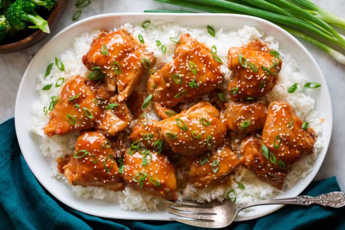 A plate of white rice topped with glazed chicken pieces garnished with sesame seeds and chopped green onions. The dish is set on a white platter with a metal fork next to it, surrounded by a bowl of broccoli and green onions in the background.