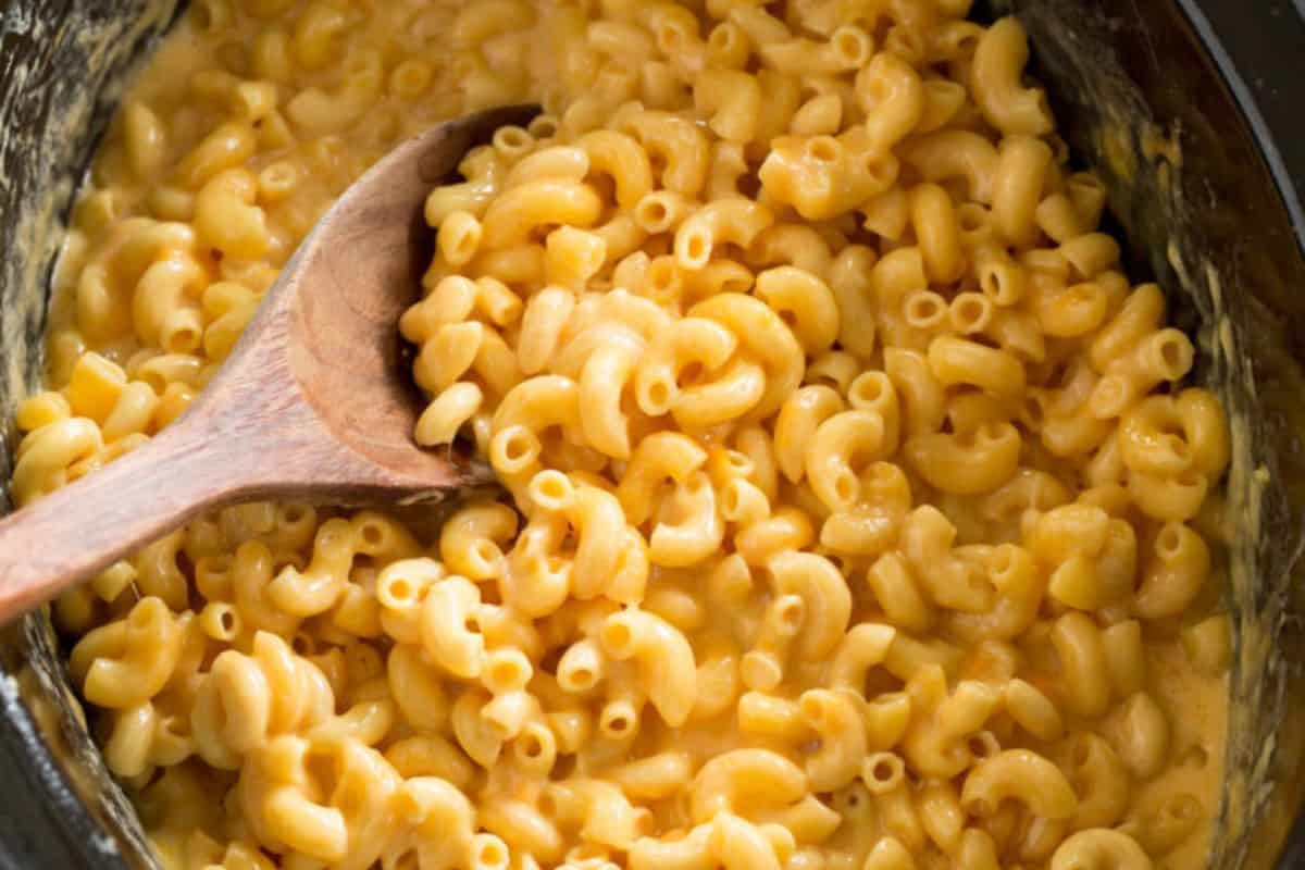A wooden spoon stirring creamy macaroni and cheese in a slow cooker.