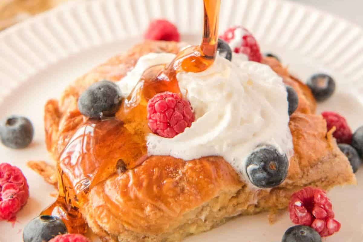 A croissant topped with whipped cream, blueberries, and raspberries, with syrup being drizzled over it. The croissant is placed on a white plate, and the vibrant colors of the berries and syrup create a visually appealing breakfast dish.