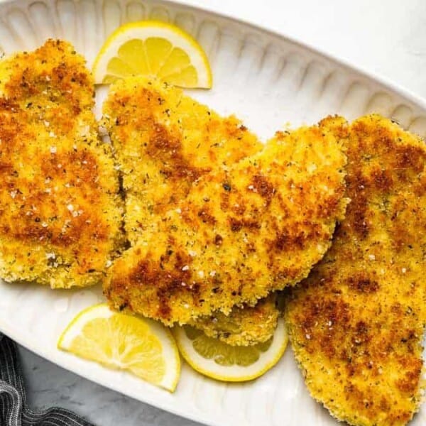A white oval plate holding four golden brown breaded chicken cutlets garnished with a sprinkling of sea salt and slices of lemon. A gray striped napkin, a small dish of lemon slices, and a bowl of greens are partially visible around the plate.