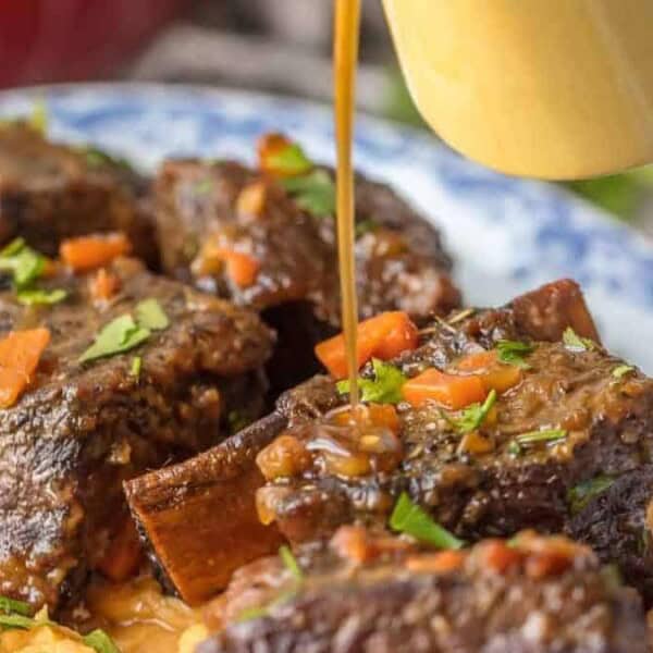 A plate of braised short ribs over mashed potatoes with diced vegetables, topped with gravy from a small pitcher.