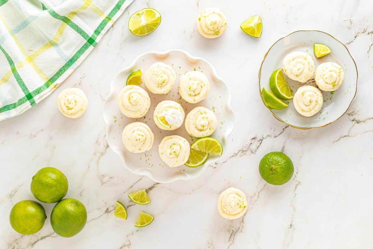 Top view of a marble countertop featuring two plates of lime cupcakes garnished with lime slices. On the countertop, there are fresh limes, lime wedges, and a green and yellow checkered towel. The cupcakes have creamy white frosting with lime zest on top.