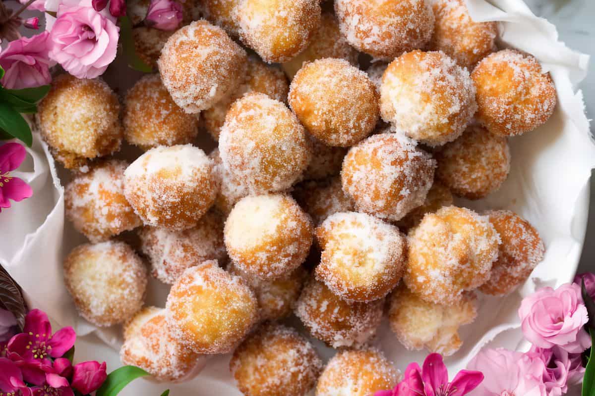 A close-up view of a pile of sugar-coated fried dough balls arranged on a white cloth, surrounded by vibrant pink flowers. The golden-brown treats are sprinkled generously with sugar, creating an inviting and appetizing display.
