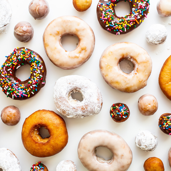 A variety of donuts and donut holes are spread out on a white background. The assortment includes glazed, chocolate with sprinkles, powdered sugar, and plain donuts. The layout is evenly spaced, displaying the donuts in an appetizing manner.