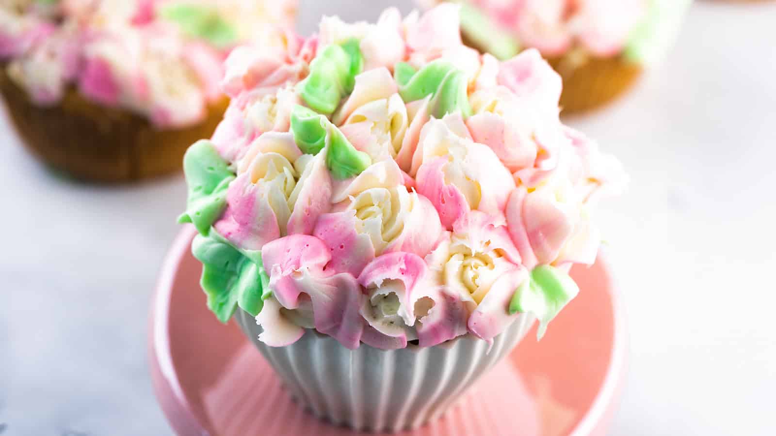 A cupcake with white and pink flower-shaped frosting and green leaves.