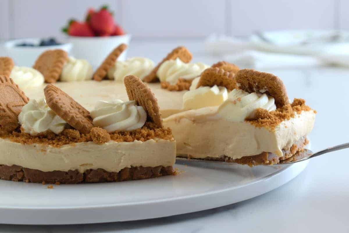 A cheesecake sits on a white plate. The cheesecake is topped with dollops of whipped cream and biscoff cookies, with crumbled cookies around the edge. A slice is being lifted with a pie server. In the background, there's a bowl of strawberries.