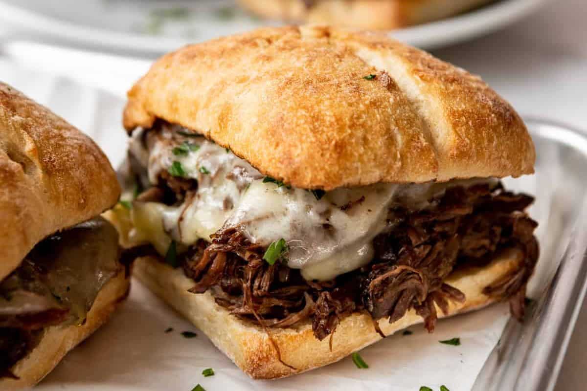 A close-up of a shredded beef sandwich with melted cheese on a golden-brown ciabatta roll. Chopped herbs are sprinkled over the cheese, and the sandwich is placed on white parchment paper on a plate. Another sandwich is partially visible in the background.
