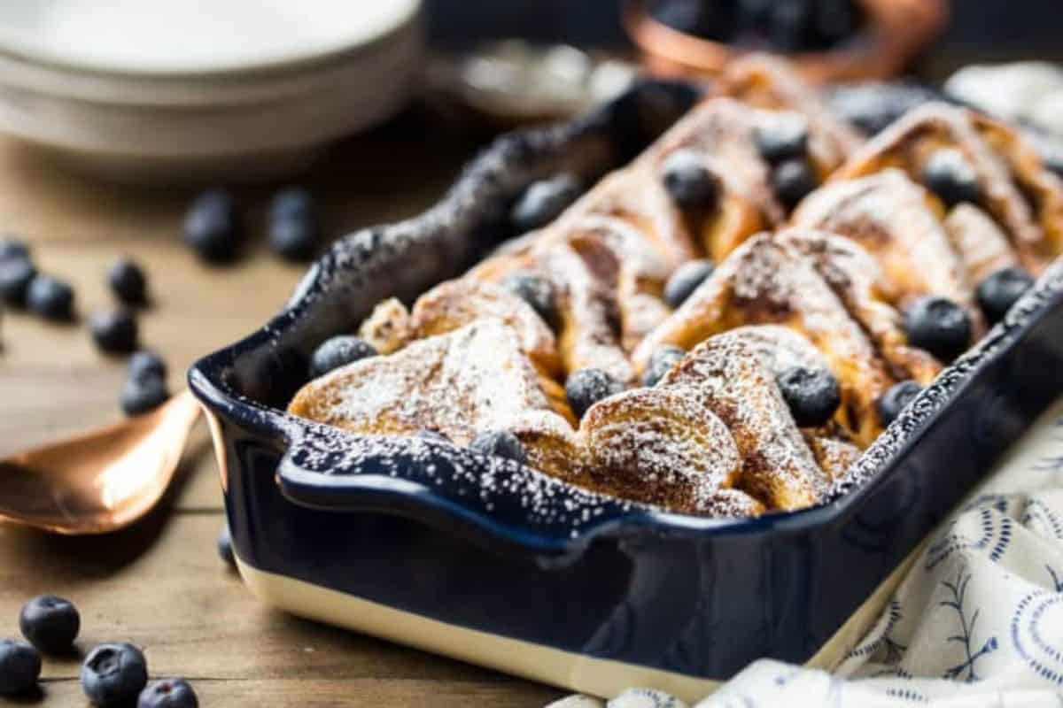 A blue ceramic dish containing freshly baked croissants dusted with powdered sugar and garnished with blueberries is on a wooden table. A copper spoon, scattered blueberries, and a stack of white plates in the background add to the cozy setting.