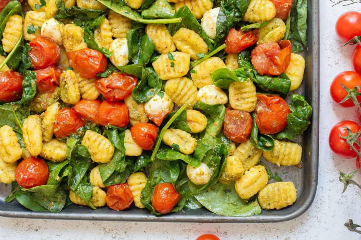 A colorful tray of baked gnocchi with roasted cherry tomatoes, fresh spinach, and mozzarella balls, seasoned with pesto. The gnocchi is lightly browned, and the tomatoes are blistered. Fresh cherry tomatoes on vine stems lie nearby on the counter.