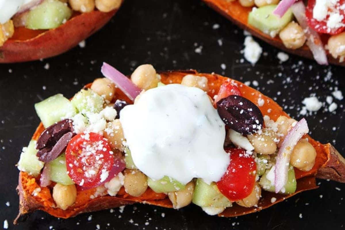 A colorful dish featuring roasted sweet potato halves topped with a mixture of chickpeas, diced cucumber, halved cherry tomatoes, red onion slices, black olives, and crumbled feta cheese. The dish is garnished with a dollop of white yogurt sauce.