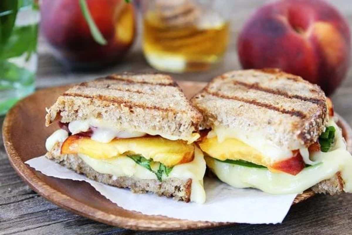 A grilled cheese sandwich with melted cheese, peach slices, and basil leaves is cut in half and placed on a wooden plate. Fresh peaches and a jar of honey are in the background, along with a green plant in a glass vase.