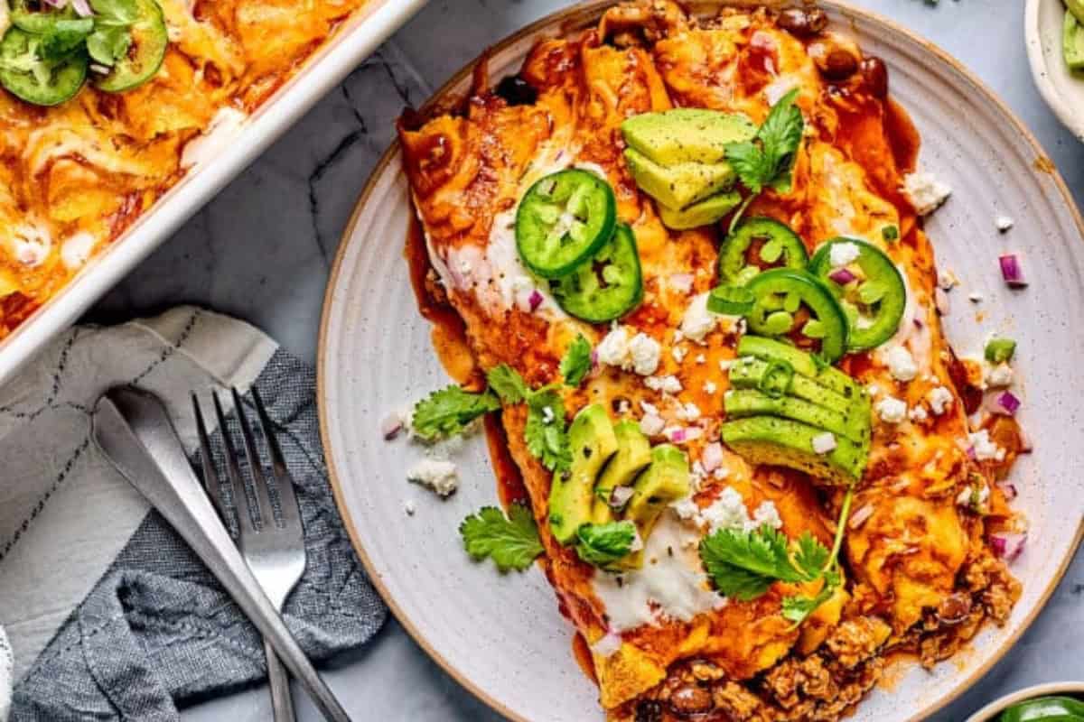 A plate of enchiladas topped with melted cheese, sliced jalapeños, avocado, cilantro, and crumbled cheese. The enchiladas are covered in red sauce and accompanied by a serving dish in the background on a marble countertop with utensils placed next to a napkin.