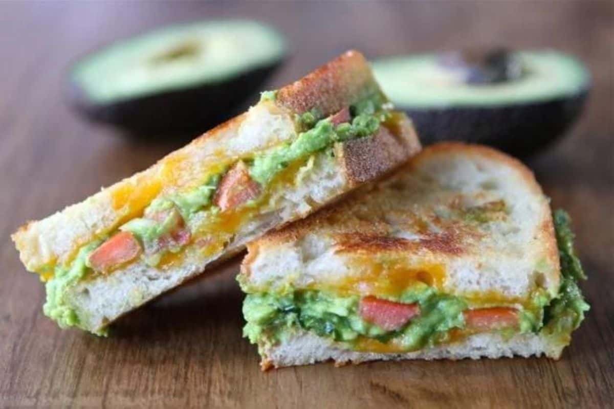 Two halves of an avocado grilled cheese sandwich are displayed on a wooden surface, with the interior showing melted cheese, avocado, and tomato. In the background, there is a halved avocado.
