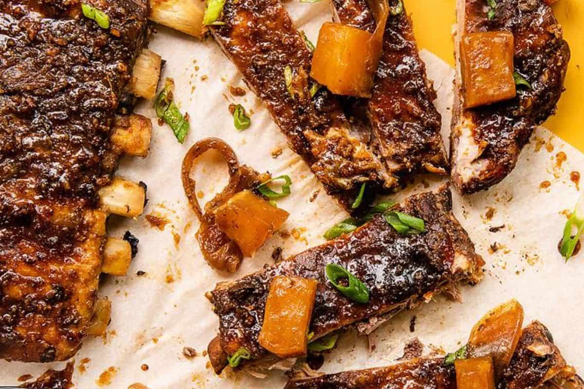 A close-up of glazed ribs on parchment paper, with a few pineapple chunks and green onion slices sprinkled on top. The ribs appear caramelized and juicy, showcasing a rich, dark glaze. The vibrant yellow background adds a pop of color to the scene.