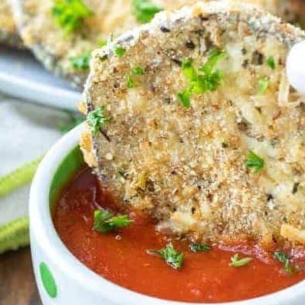 A hand dips a breaded, baked eggplant slice garnished with parsley into a bowl of marinara sauce.