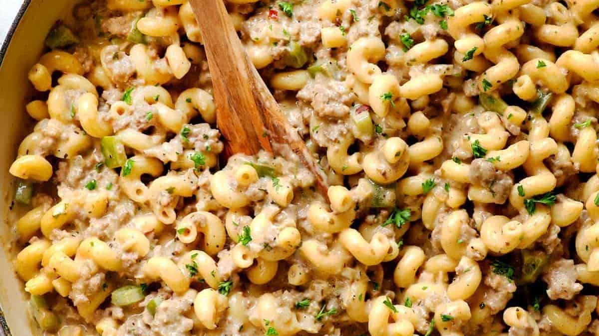 A close-up of creamy macaroni pasta with ground beef and chopped green peppers, garnished with parsley and stirred with a wooden spoon.