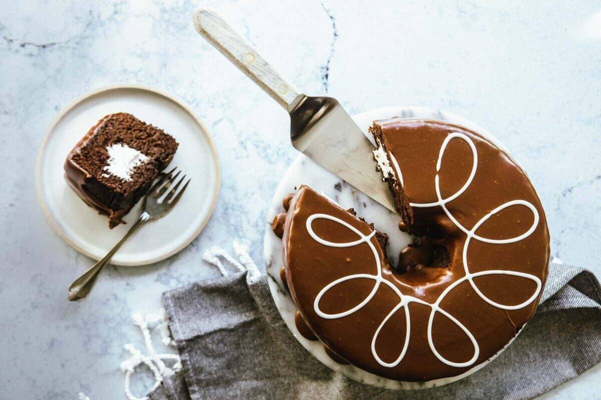 A chocolate cake with white icing swirl on a marble platter, one slice on a plate with a fork, set on a white surface with a gray napkin.