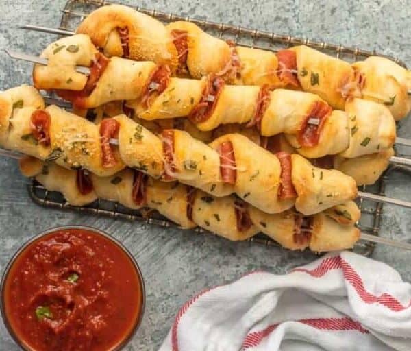 Four skewers of twisted bread dough with pepperoni and herbs on a wire rack beside a bowl of marinara and a white towel with red stripe.