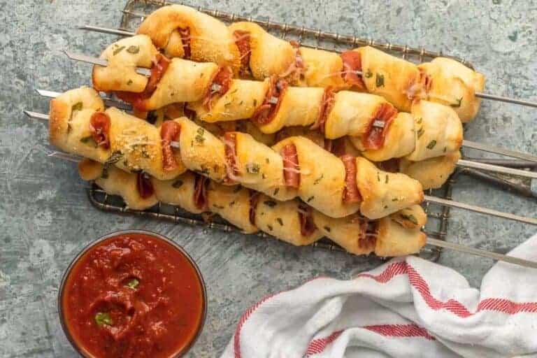 Four skewers of twisted bread dough with pepperoni and herbs on a wire rack beside a bowl of marinara and a white towel with red stripe.