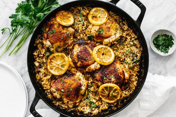 A skillet with browned chicken thighs on seasoned rice, topped with lemon slices and herbs. Parsley and chopped herbs sit beside it.
