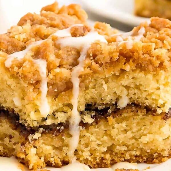 A close-up of a slice of crumb-topped coffee cake with a cinnamon layer, icing drizzle, and a fork on a white plate.