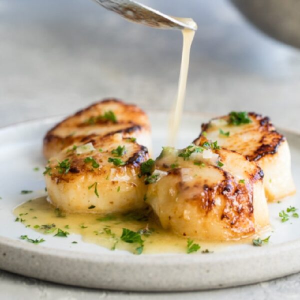 Four seared scallops on a white plate are topped with chopped herbs, and a light sauce is being poured over them.