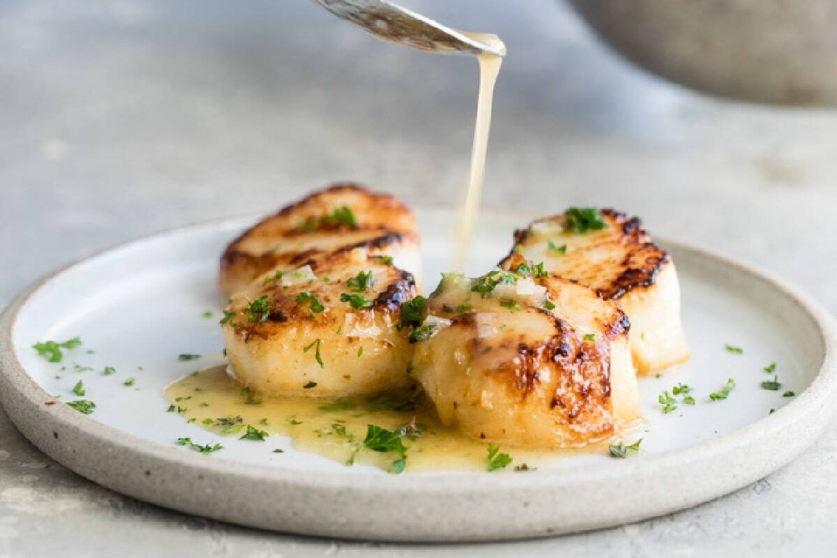 Four seared scallops on a white plate are topped with chopped herbs, and a light sauce is being poured over them.