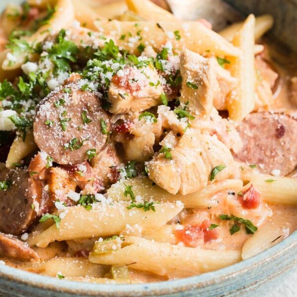A bowl of creamy penne pasta with sliced sausage, chicken pieces, diced tomatoes, parsley, and grated cheese on top.