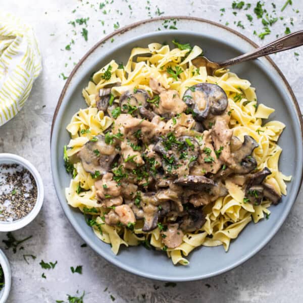 A bowl of egg noodles with creamy mushroom and beef stroganoff, topped with parsley, sits beside a fork, seasonings, and a napkin.