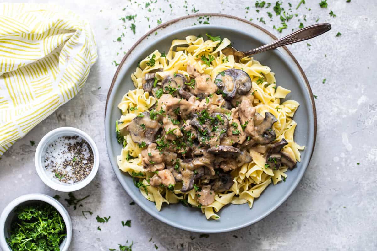 A bowl of egg noodles with creamy mushroom and beef stroganoff, topped with parsley, sits beside a fork, seasonings, and a napkin.