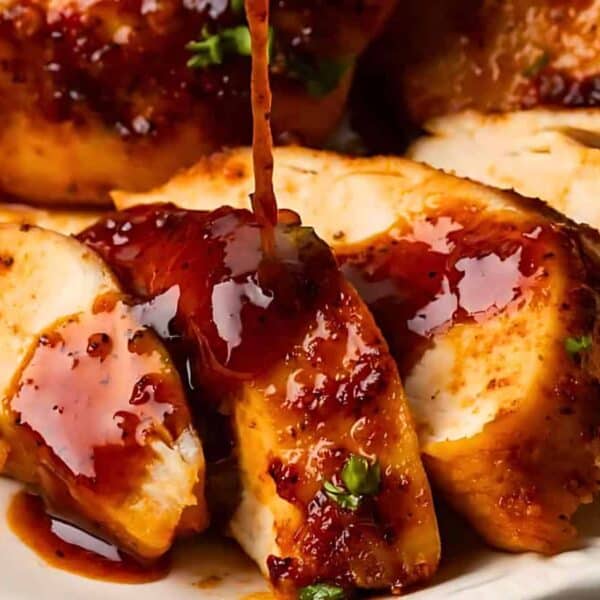 Sliced cooked chicken breast on a plate with glossy reddish-brown sauce being poured over the top.
