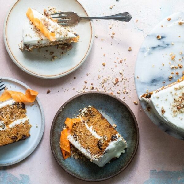 Three slices of carrot cake with cream cheese frosting and carrot ribbons on plates, beside a partially sliced cake on a marble stand.