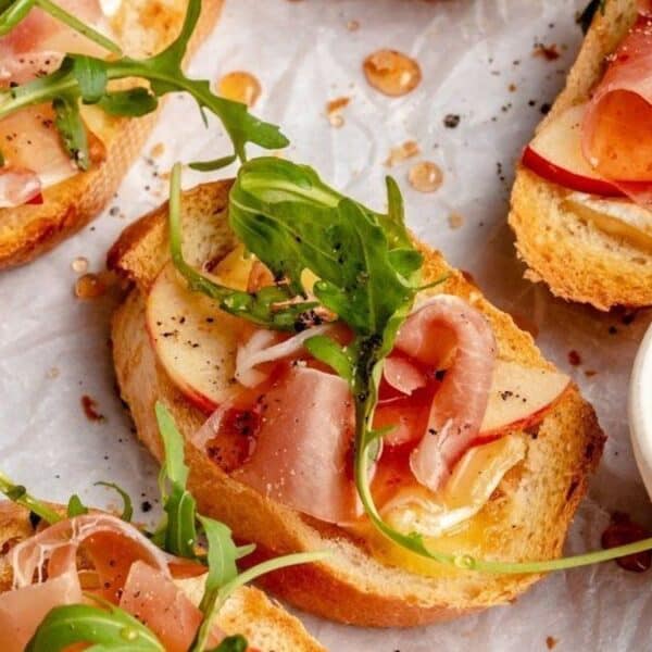 Slices of toasted bread topped with prosciutto, apple slices, arugula, and black pepper are arranged on parchment paper beside a small bowl of honey.